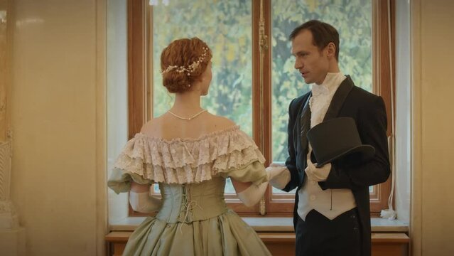 Back view medium shot of young Caucasian aristocratic couple in medieval outfits standing by window in classic ballroom and having conversation