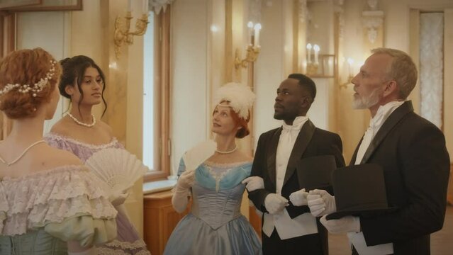 Medium slowmo of elegant ethnically diverse ladies and gentlemen wearing luxurious ball outfits and suits having conversation while standing in classic style corridor of aristocratic ball hall