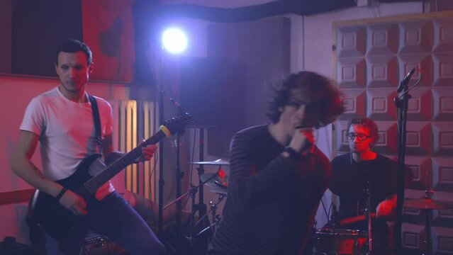 The lead singer of the rock band sings aggressively into the microphone, gestures emotionally and moves to the beat of the music.  The vocalist, drummer and guitarist perform the song in rehearsal.