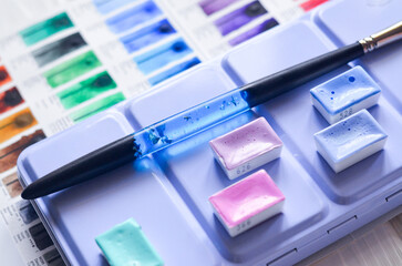 palette of watercolor paints lies on a wooden white table, watercolor godets, pastel colors, brush...