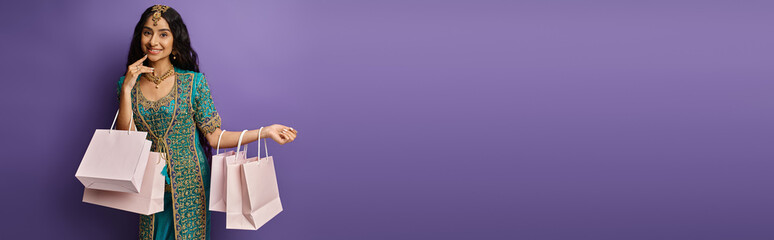 happy beautiful indian woman in blue sari posing with shopping bags on purple background, banner