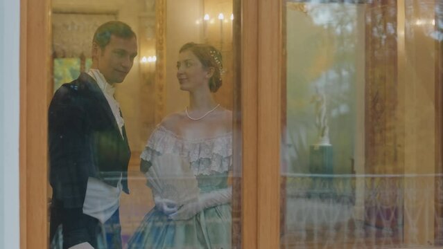 Medium through window slowmo of young Caucasian couple wearing Regency style dresses looking at window and having conversation while spending time at debutante ball together