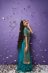 attractive cheerful indian woman in blue sari posing under confetti rain on purple background