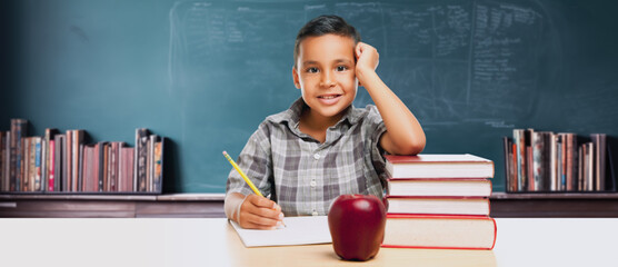 Happy Young Hispanic School Boy At Desk with Books and Apple In Front of Chalkboard.