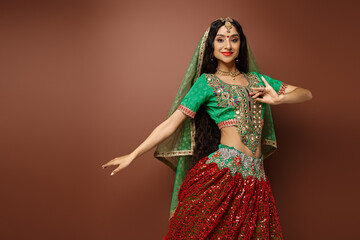 cheerful young indian woman in green choli with bindi dot gesturing while dancing looking at camera