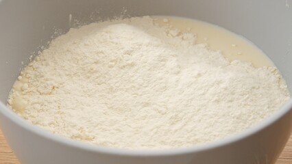 Flour pours into white bowl. A fine stream of white flour is added to the contents of a bowl,...