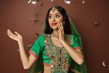 Obraz premium cheerful pretty indian woman in traditional clothing posing on brown backdrop under confetti rain
