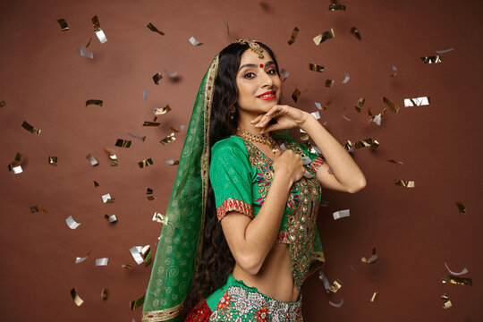 Pretty Indian Woman With Bindi Dot And Green Veil Posing Under Confetti Rain With Hand Under Chin