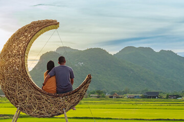 Happy couple sit on crescent moon chair made of rattan for relaxation in paddy field with beautiful scenic in evening. Decorative wooden moon furniture as sitting chair for viewpoint in rice field.