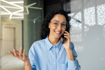 Young beautiful woman smiling and looking at camera, talking on the phone, businesswoman at workplace inside office, worker in glasses shirt and curly hair.