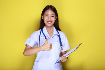 A young woman wearing a doctor's uniform poses with a thumbs up and hangs a medical stethoscope,...