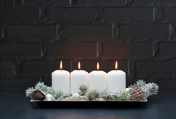 Four white burning candles with Christmas decorations in front of a black brick wall with copy...