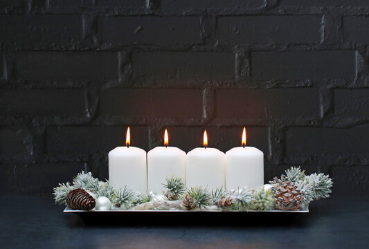 Four white burning candles with Christmas decorations in front of a black brick wall with copy space.