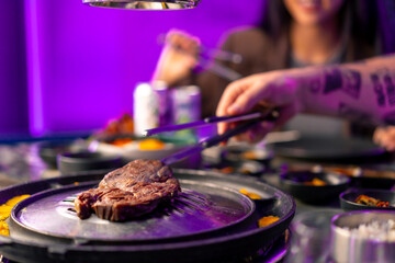 close-up of a hand with tongs picking up a juicy raw piece of meat to put on the grill in a Korean...