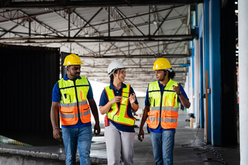 A team of warehouse workers in uniforms wearing yellow hard hats chats in a large logistics...