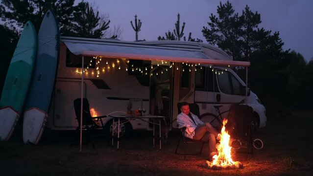 A man is sitting by the campfire in the evening, wrapped in a blanket. Evening rest near a mobile home