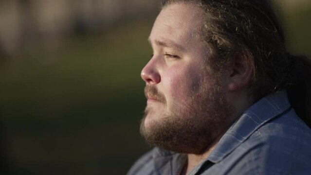 One contemplative overweight young man sitting outdoors in mental reflection. Pensive thoughtful expression of a chubby male caucasian person during sunset at park