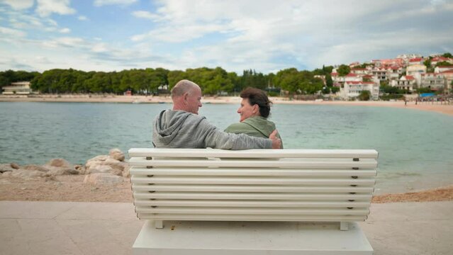 Retirement holiday, joyful elderly woman and man are sitting in an embrace on a bench near sea coast during a tourist trip