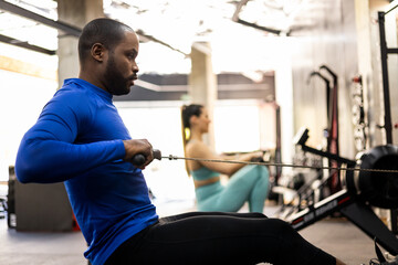 A couple of athletes of different ethnicities are working their bodies with a magnetic air rowing machine. The African adult is in the foreground performing a back exercise with the row. - obrazy, fototapety, plakaty