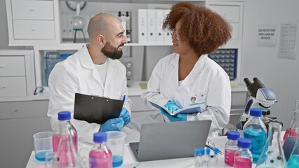 Two smiling scientists engage in online conference, reading a book and writing on clipboard in their lab while working on a medical research