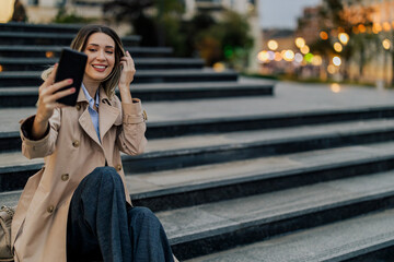 Fototapeta premium Woman sitting on a stairs on a city street and taking selfie with a smartphone