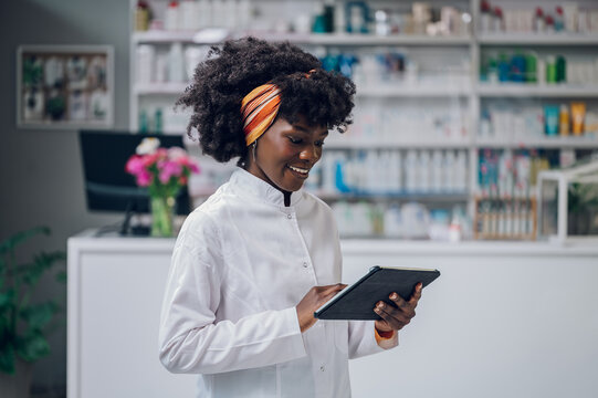 A cheerful young multiracial druggist is standing in a drugstore and smiling at the tablet while scrolling on it.