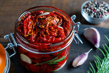 Sun-dried red tomatoes with garlic, green rosemary, olive oil and spices in a glass jar on a wooden...