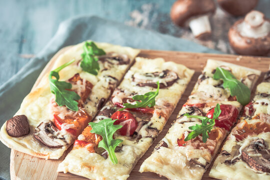 Freshly baked flammkuchen, traditional french tarte flambee or german pizza in a vegetarian recipe with mushrooms, cream cheese, tomatoes and arugula, on  light greenwooden board and table
