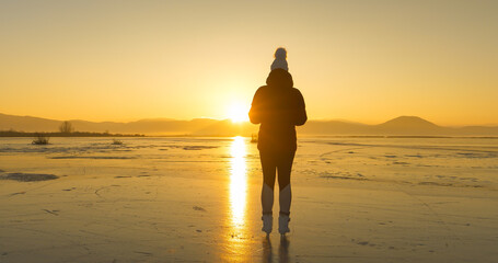 LENS FLARE, SILHOUETTE: Ice skater stands on frozen lake and admires setting sun