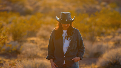 A fearless cowgirl, adorned in a rugged cowboy hat, strides through the harsh desert landscape of...