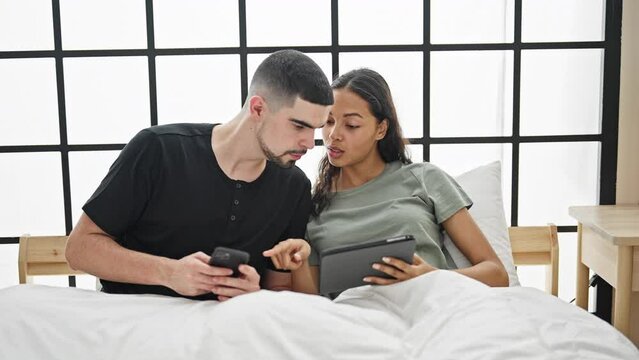 Beautiful couple sitting together on a cozy bed in the bedroom, immersed in their smartphone and touchpad, basking in the relaxed morning at home