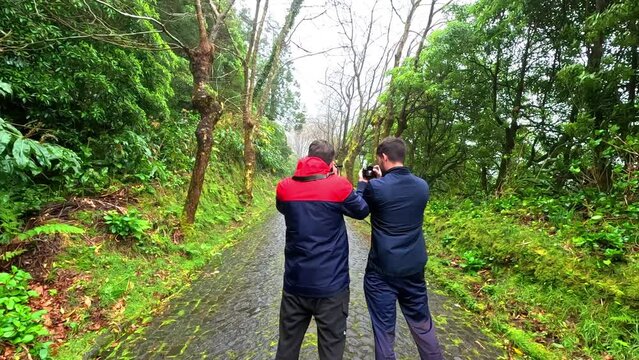 Two men photographer taking pictures in the middle of a path surrounded of green trees and moss and going into the fog, in a rainy day with a lot of moisture