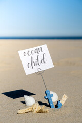 Obraz premium OCEAN CHILD text on paper greeting card in anchor paper holder and starfish seashell summer vacation decor. Sandy beach sun coast. Holiday concept postcard. Getting away Travel