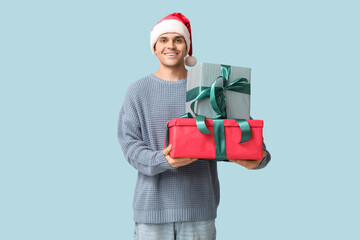 Handsome young man in Santa hat holding Christmas gifts on blue background