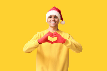 Handsome young man in Santa hat making heart with hands on yellow background