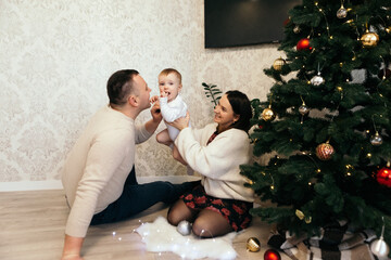 Family mother father little boy cute child playful toddler sitting near Christmas Tree playing New Year decorations toys balls decorated interior holiday atmosphere gifts presents garlands
