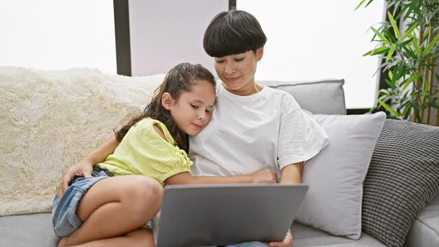 Mother and daughter share a loving kiss on the sofa at home while happily using laptop, engrossed in a feel-good family movie