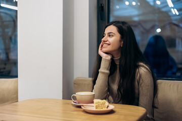 Pretty smiling young woman sitting in cafe, drink cup of coffee hot beverage, eating piece of...