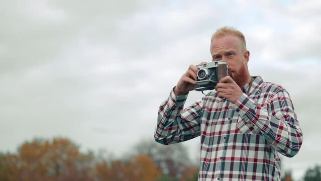 Male traveller in checkered T-shirt walking in a park on a gloomy day, looking for stunning point outdoors, taking pictures. Blurred view of autumn trees. High quality 4k footage