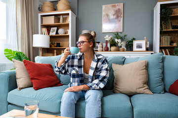 Obraz premium Young smiling woman sitting on sofa and looking up while drinking hot tea or coffee. Young brunette woman thinking at home in a leisure time. Happy girl relaxing at home on a bright winter morning.