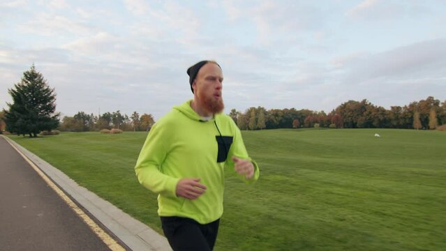 Young sportsman in black bini hat with red beard exercising in autumn park, running with mouth open, looking under his feet. Training in the fresh air. High quality 4k footage