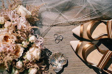 Wedding rings, bride shoes and a bouquet lie on a wooden table. Top view
