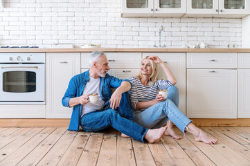 Smiling mid aged woman and man sit on wooden floor in cozy kitchen