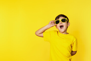 Excited young boy in vibrant yellow lifting sunglasses on a matching background, expressing...