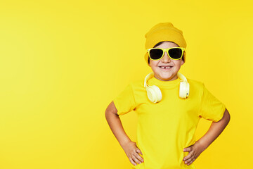 Happy young boy in yellow outfit and shades posing confidently with headphones, vibrant yellow...