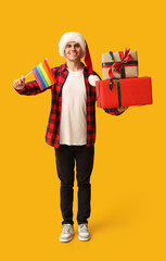 Young man in Santa hat with LGBT flag and Christmas gifts on yellow background