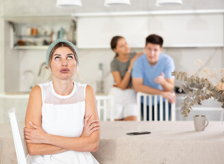 Emotionally tense frustrated woman sitting at table in home kitchen against blurred background of dissatisfied daughter standing with husband behind. Concept of family discord