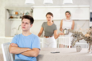 Obraz premium Upset young guy sitting at kitchen table on background of dissatisfied wife standing with mother-in-law behind. Family conflicts and communication problems concept