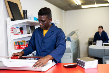 Naklejka na ściany i meble Serious middle-aged African American male in a blue uniform using paper cutter in the printer house