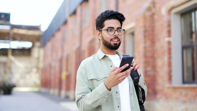 A handsome student with a backpack uses a smartphone while walking around the campus space near the university building. Happy smiling male chatting online, browsing social media or reading messages